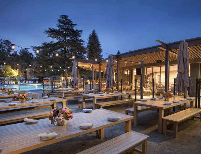 Outdoor dining area with wooden tables, benches, string lights, and floral arrangements. A pool and trees are visible in the background.